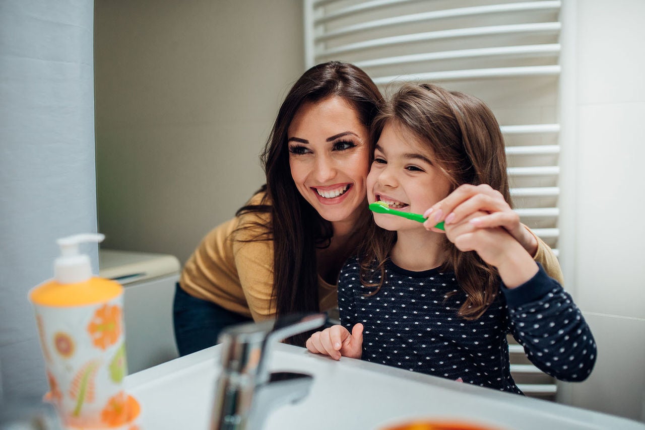 Frau und 7-jähriges Mädchen lächeln in den Spiegel, während sie sich die Zähne putzen.